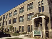 Exterior view of a three-story brick building with multiple windows and a small covered entrance. A sign near the entrance reads 'John Manley House'. There are a few cars parked along the sidewalk and a street lamp visible on the left side.