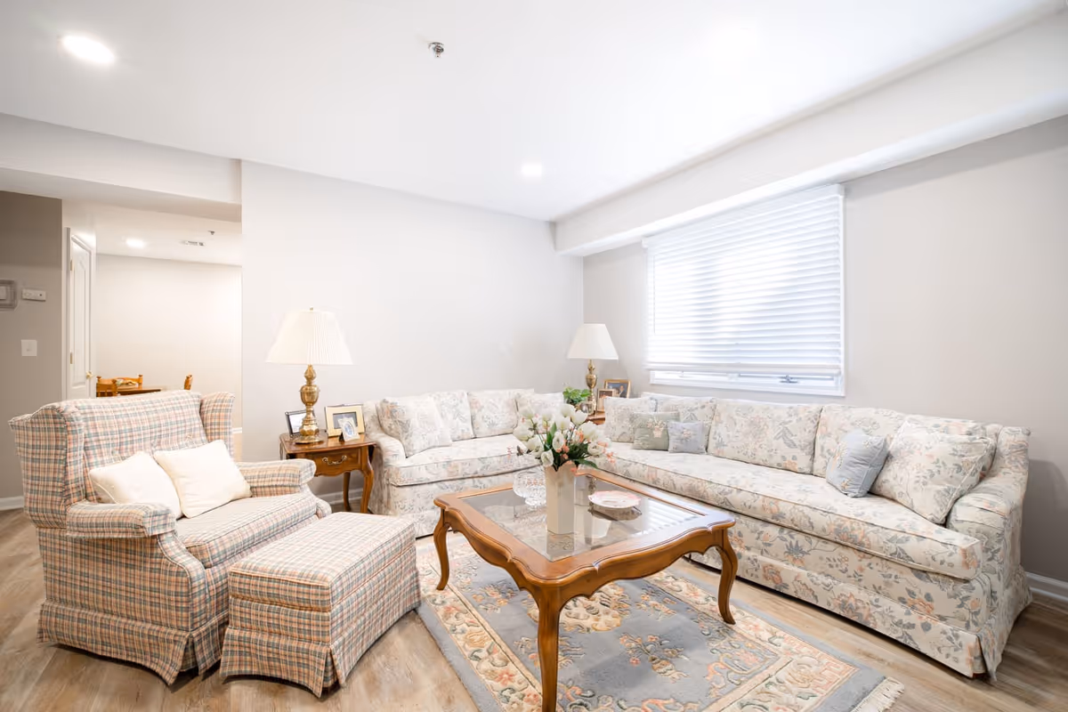 A bright and cozy living room with a floral-patterned sofa, a matching loveseat, and a plaid armchair with an ottoman. There is a wooden coffee table with a glass top and a vase of flowers in the center. Two table lamps with white shades sit on side tables next to the sofas. A large window with white blinds allows natural light to fill the room. The floor is wood with a decorative area rug.