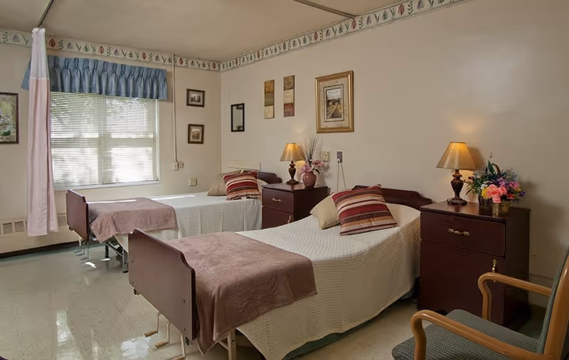 A room with two single beds each covered with white bedspreads and brown blankets at the foot. Each bed has a striped pillow and a beige pillow. Between the beds are two dark wooden nightstands with table lamps, a vase with flowers, and framed pictures on the wall above. A window with blinds and a blue valance is on the left side, and a curtain hangs partially in front of the window. There is a wooden chair with green upholstery in the foreground.