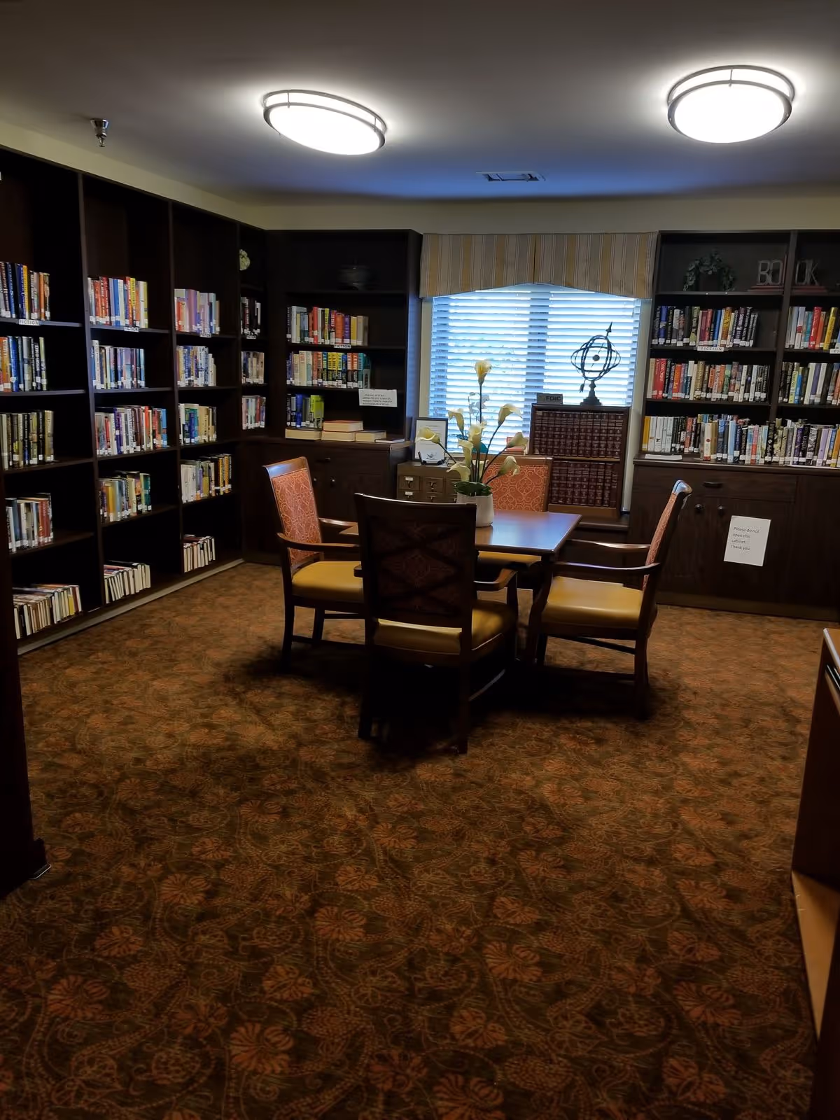 Cozy library-style room with bookshelves lining the walls and a table with four chairs beneath ceiling lights.
