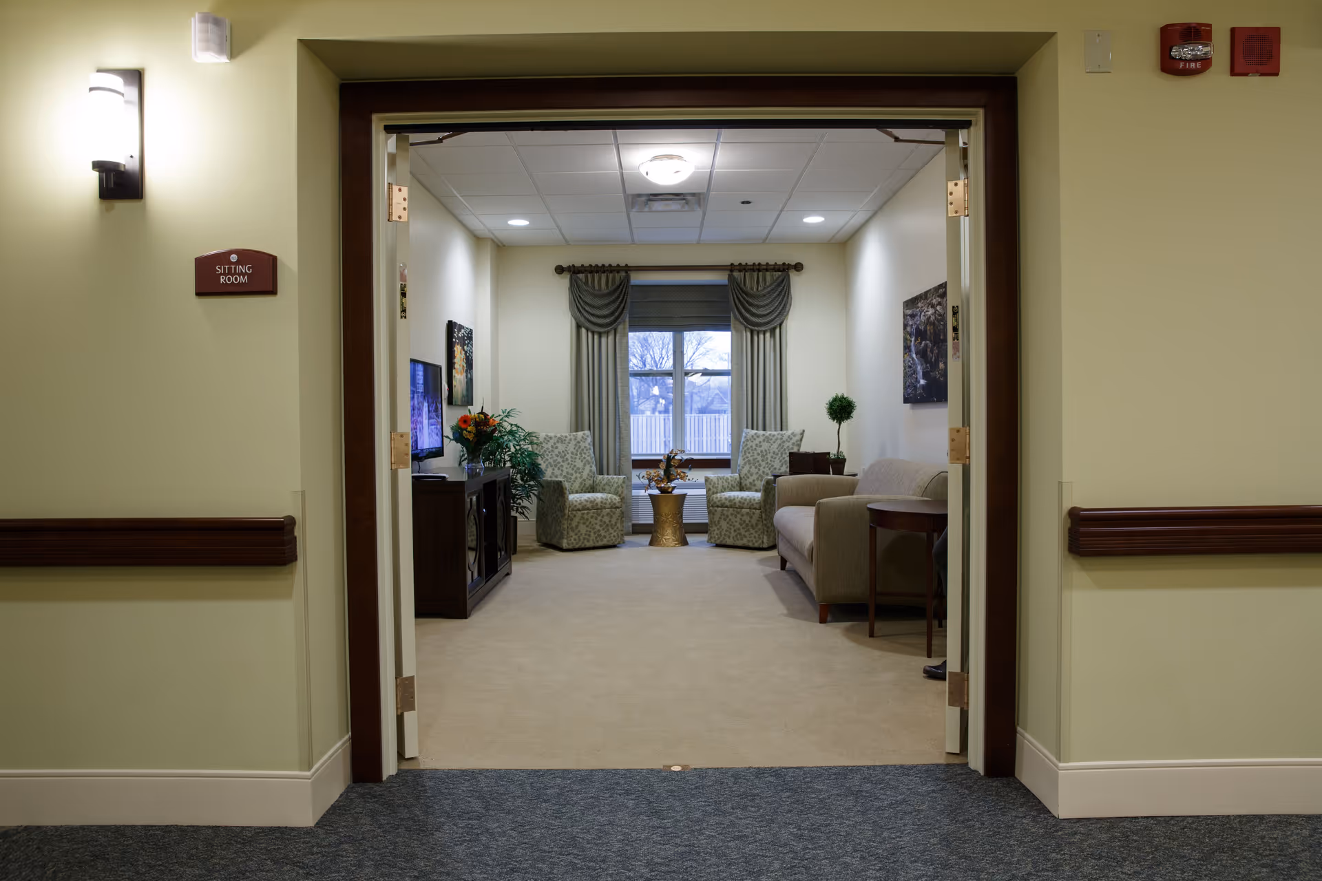 Open double doors leading into a cozy sitting room with armchairs, a sofa, a TV cabinet, and a window with drapes.