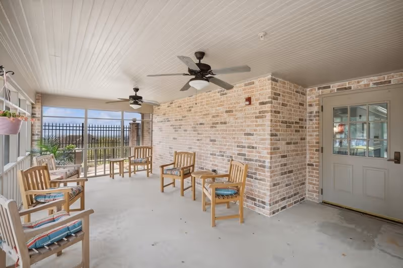 A covered outdoor patio area with brick walls and a white ceiling featuring two ceiling fans. The space is furnished with wooden chairs with cushions and small wooden tables. The patio is enclosed with large windows and a black metal fence is visible outside. There are potted plants hanging and placed near the windows.