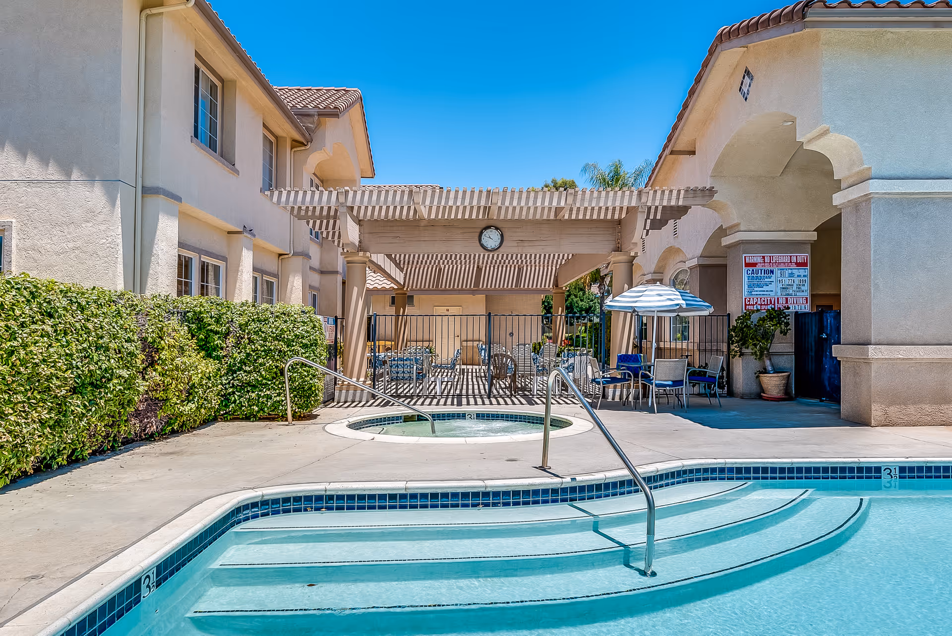 Outdoor pool and hot tub area with steps, metal handrails, a pergola-covered seating area and umbrellas in front of a beige senior living building under a clear blue sky.