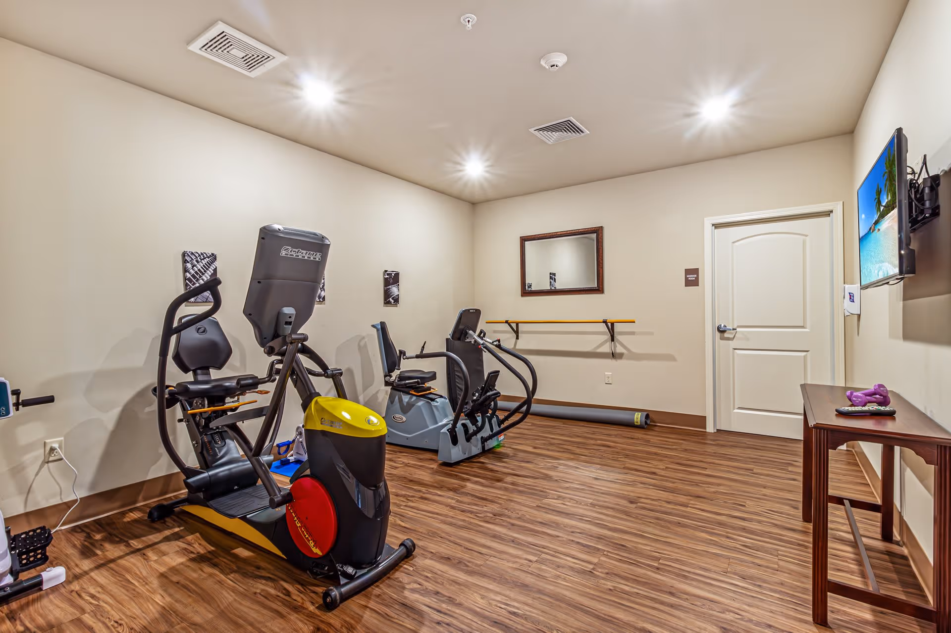 A small exercise room with two recumbent exercise bikes, a wall-mounted mirror, a ballet barre, a wall-mounted television displaying a beach scene, and a wooden table with purple dumbbells and a remote control. The room has wood flooring, beige walls, and recessed ceiling lights.