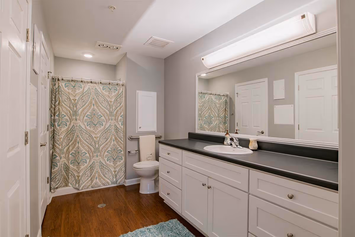 A clean and modern bathroom with a large mirror above a black countertop and white cabinets. There is a toilet with a towel hanging on a grab bar next to it, and a shower with a patterned curtain. The floor is wooden, and the walls are painted light gray.