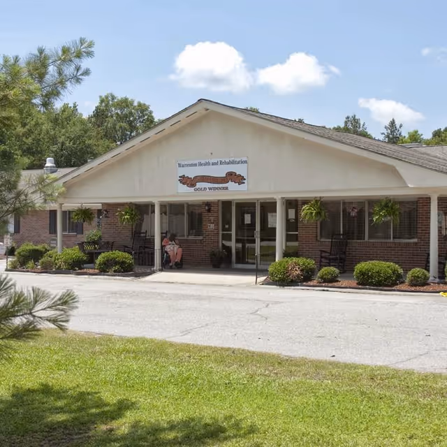 Front entrance of a one-story brick health and rehabilitation facility with a covered porch, hanging plants, shrubs, and people seated near the doorway.