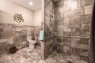 Spacious accessible bathroom with extensive gray tile, a large walk-in shower with grab bars, a toilet, towel, and decorative wall art.