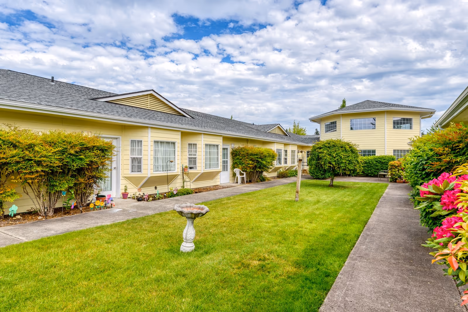 A well-maintained outdoor courtyard area of a senior living facility with green grass, a birdbath, trimmed bushes, and a paved walkway. The buildings surrounding the courtyard are painted yellow with white trim, featuring multiple windows and doors. The sky is partly cloudy.