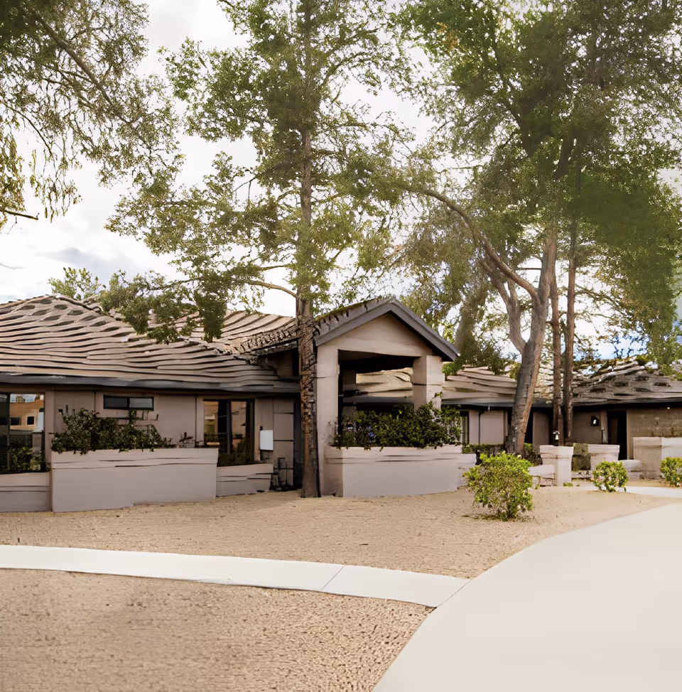 Exterior view of a single-story building with a covered entrance, surrounded by trees and desert landscaping with gravel and small bushes.