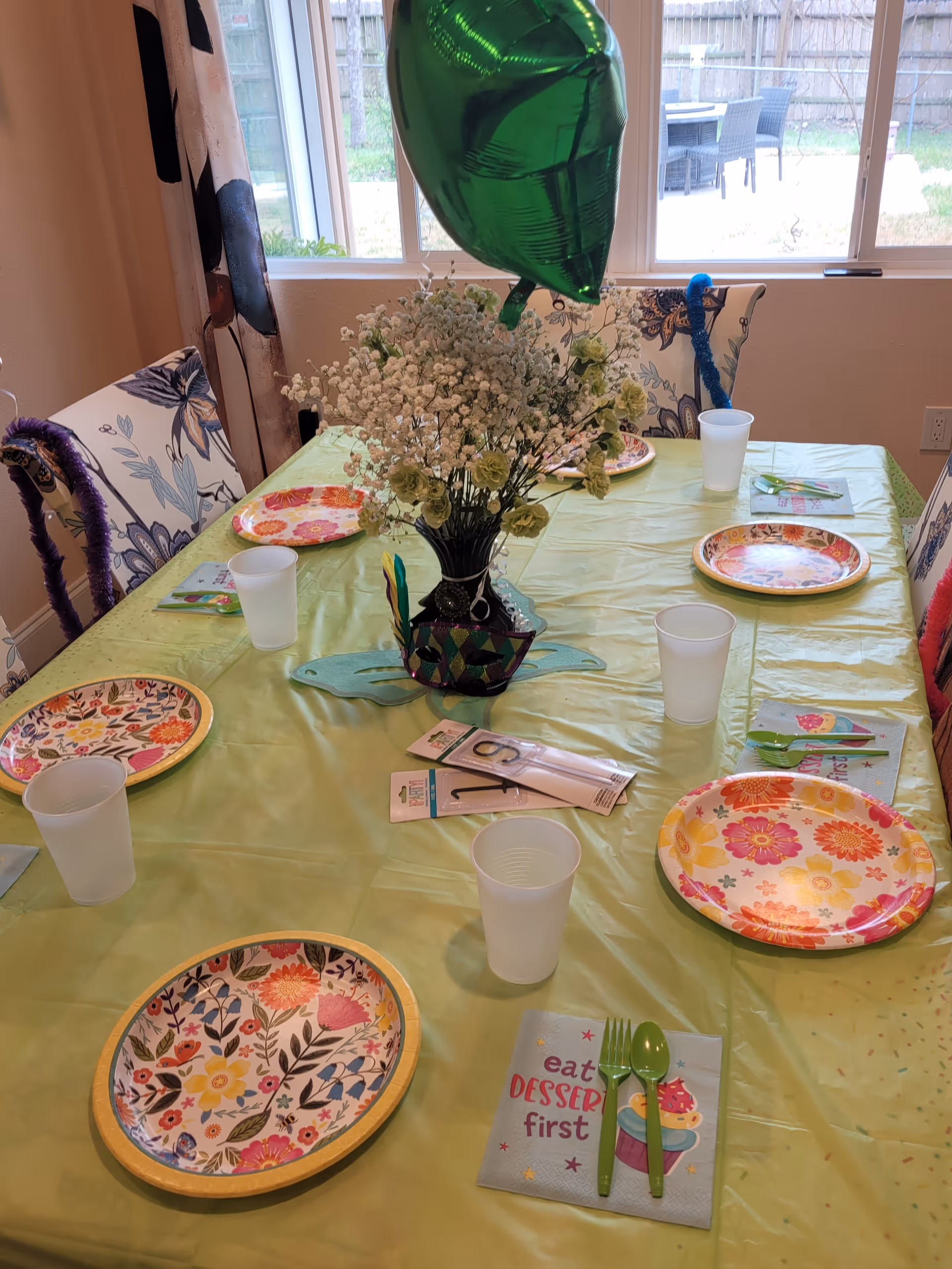 A dining table set for a celebration with floral-patterned paper plates, plastic cups, green plastic utensils on napkins that say 'eat DESSERT first', and a centerpiece of white flowers in a decorative vase. There is a green balloon attached to the centerpiece. The table is covered with a light green tablecloth and surrounded by chairs with floral upholstery. A window in the background shows an outdoor patio area.
