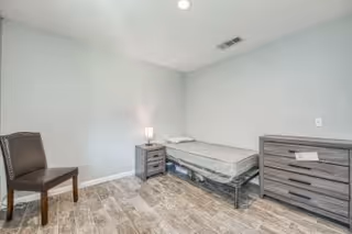A simple bedroom with a single bed, a nightstand with a lamp, a wooden chair, and a chest of drawers. The room has light-colored walls and wood-look tile flooring.