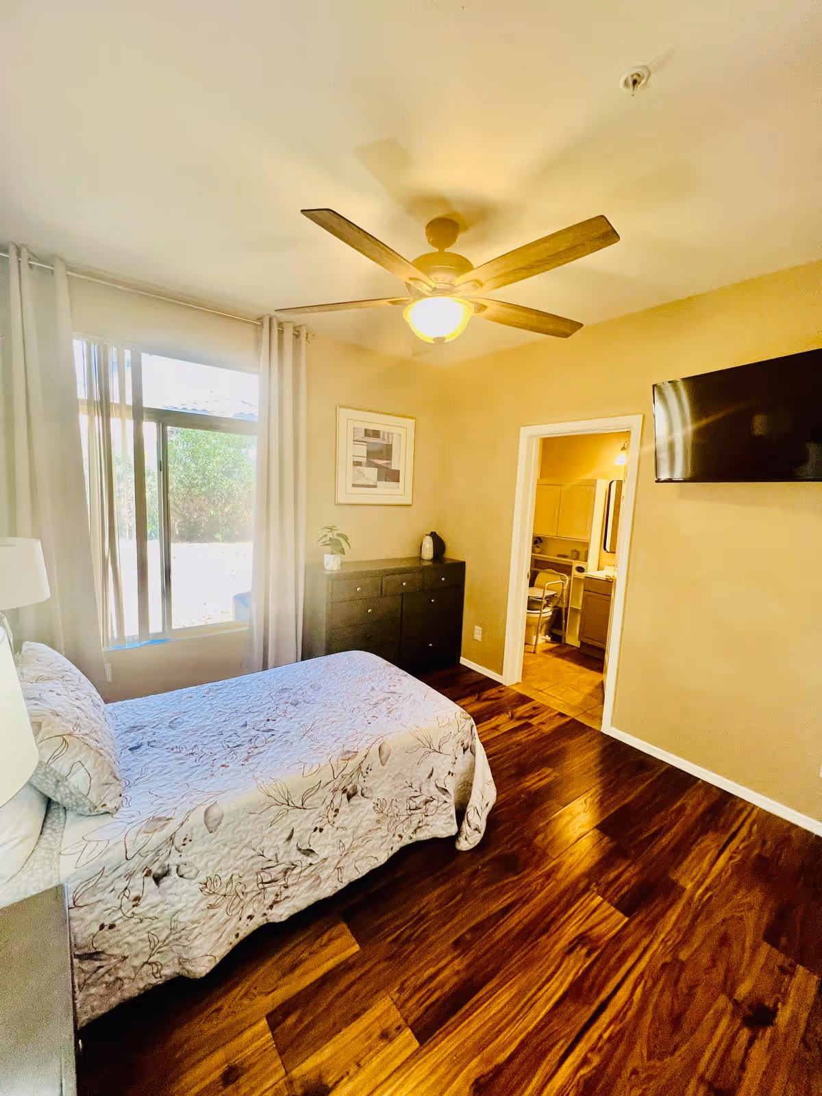 A bedroom with a bed covered in a floral quilt, a window with white curtains, a ceiling fan with a light, a black dresser with a plant and decorative item on top, a wall-mounted TV, and an open door leading to a bathroom.