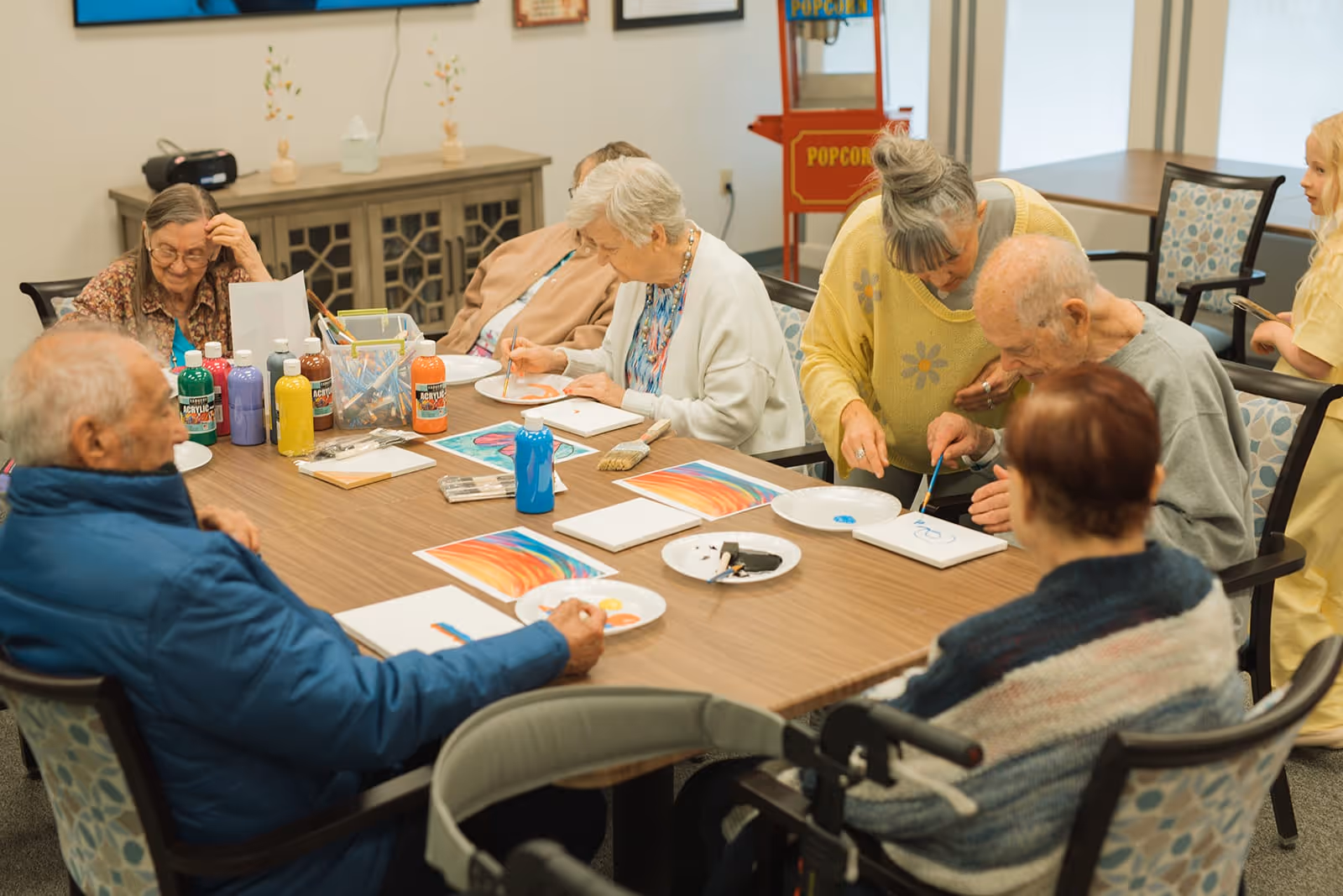 A group of elderly people sitting around a table engaged in a painting activity with various colors of acrylic paint and brushes. A caregiver or instructor is assisting one of the elderly men with his painting. The room has a cabinet in the background and a popcorn machine near the window.