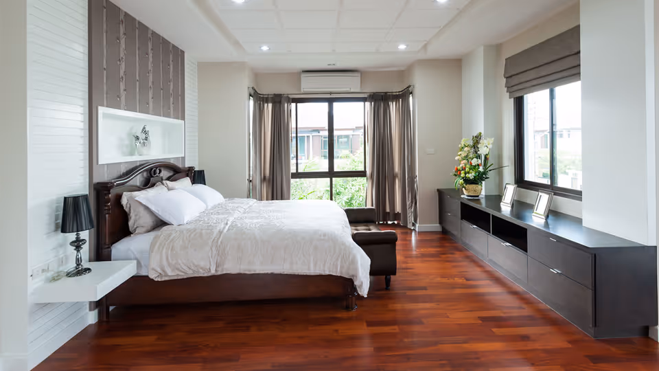 A bright and spacious bedroom with a large wooden bed featuring white bedding and pillows. The room has wooden flooring, two windows with brown curtains and blinds, a long dark wooden cabinet with decorative items and photo frames, and a small bench at the foot of the bed. The walls are light-colored, and there is a modern lamp on a floating white nightstand beside the bed.