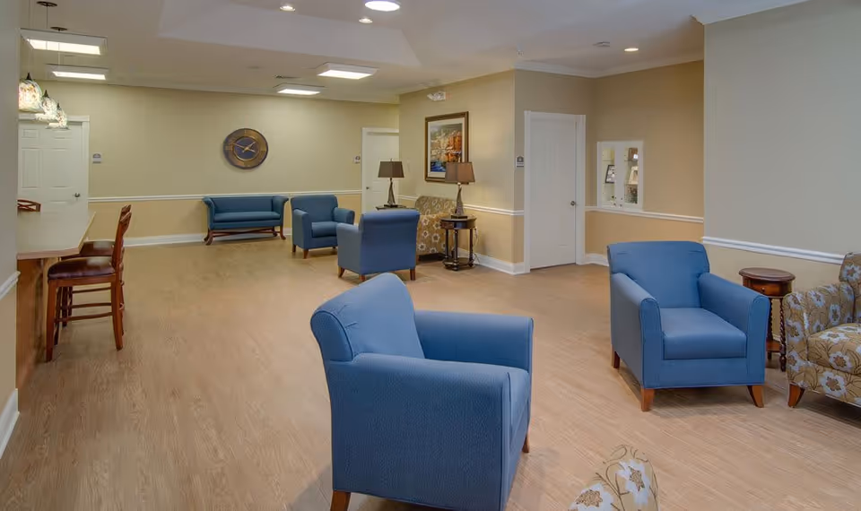 A spacious senior living facility common area with light wood flooring and beige walls. The room features several blue upholstered armchairs, a floral patterned sofa, wooden side tables with lamps, a wall clock, and framed artwork. There is a counter with bar stools on the left side and two white doors in the background.