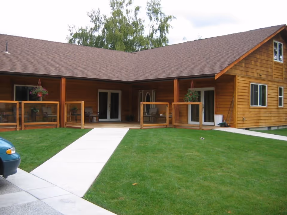 Exterior view of a single-story wooden building with a brown shingled roof, a concrete walkway leading to a covered porch area with glass-paneled wooden railings, hanging flower baskets, and patio chairs. Green grass surrounds the walkway and building.