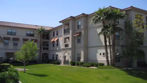 Three-story beige senior living building with arches, balconies, and palm trees in a green courtyard.