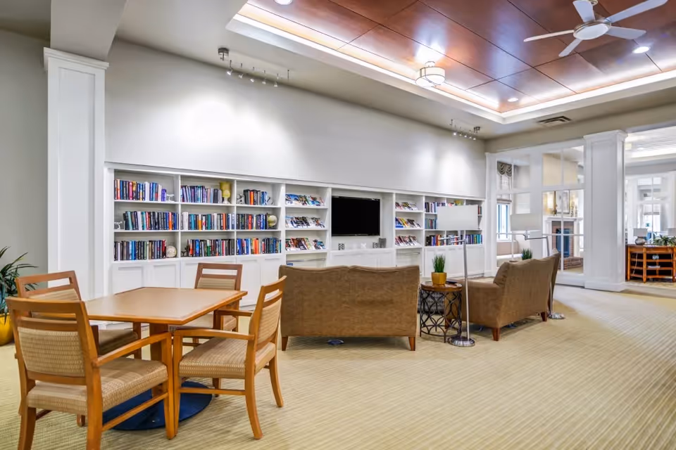 A spacious and well-lit common area featuring a wooden table with four chairs in the foreground, two brown upholstered armchairs and a matching loveseat facing a built-in white bookshelf filled with books and magazines. A flat-screen TV is mounted in the center of the bookshelf. The ceiling has wooden paneling with recessed lighting and a ceiling fan. The room has beige carpeting and large windows in the background allowing natural light.