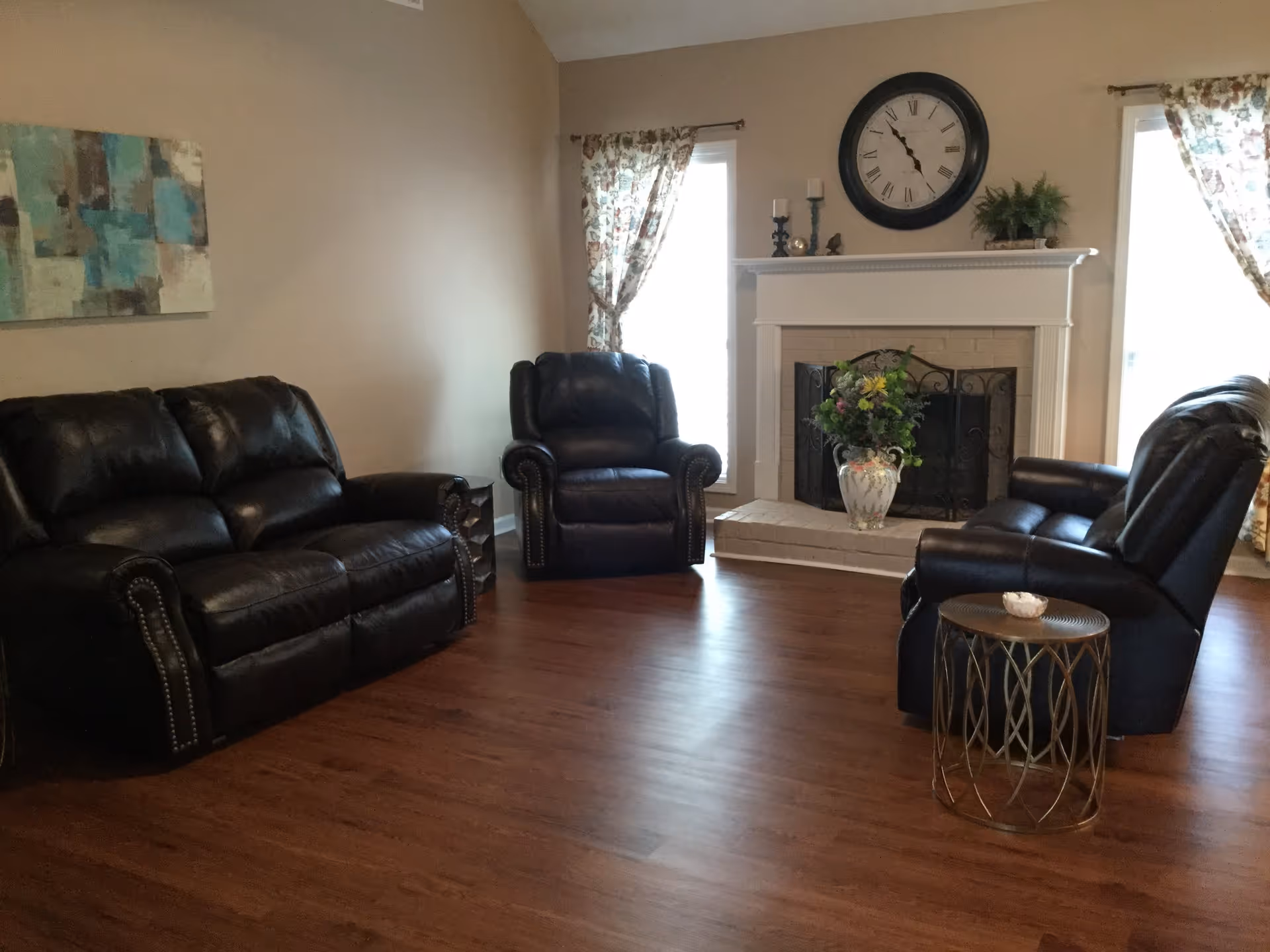 A cozy living room with dark leather sofas and armchairs arranged around a white brick fireplace. The fireplace mantel holds decorative items including candles and a plant, with a large round clock mounted above it. Two windows with floral curtains flank the fireplace, and a small round side table is placed next to one of the armchairs. The room has wooden flooring and beige walls.