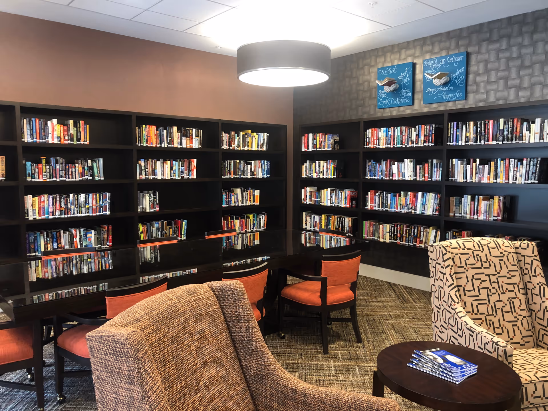 Community library/reading room with wall bookshelves, a long glossy table with orange chairs, and upholstered armchairs.