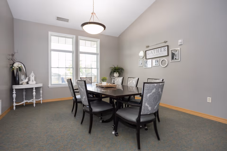 A dining room with a rectangular dark wooden table surrounded by six upholstered chairs with patterned backs. The room has light gray walls, a carpeted floor, and two large windows letting in natural light. A white side table with decorative items is placed against one wall, and a gallery wall with framed pictures and a sign that reads 'GATHER' is visible on another wall. A ceiling light fixture hangs above the table.