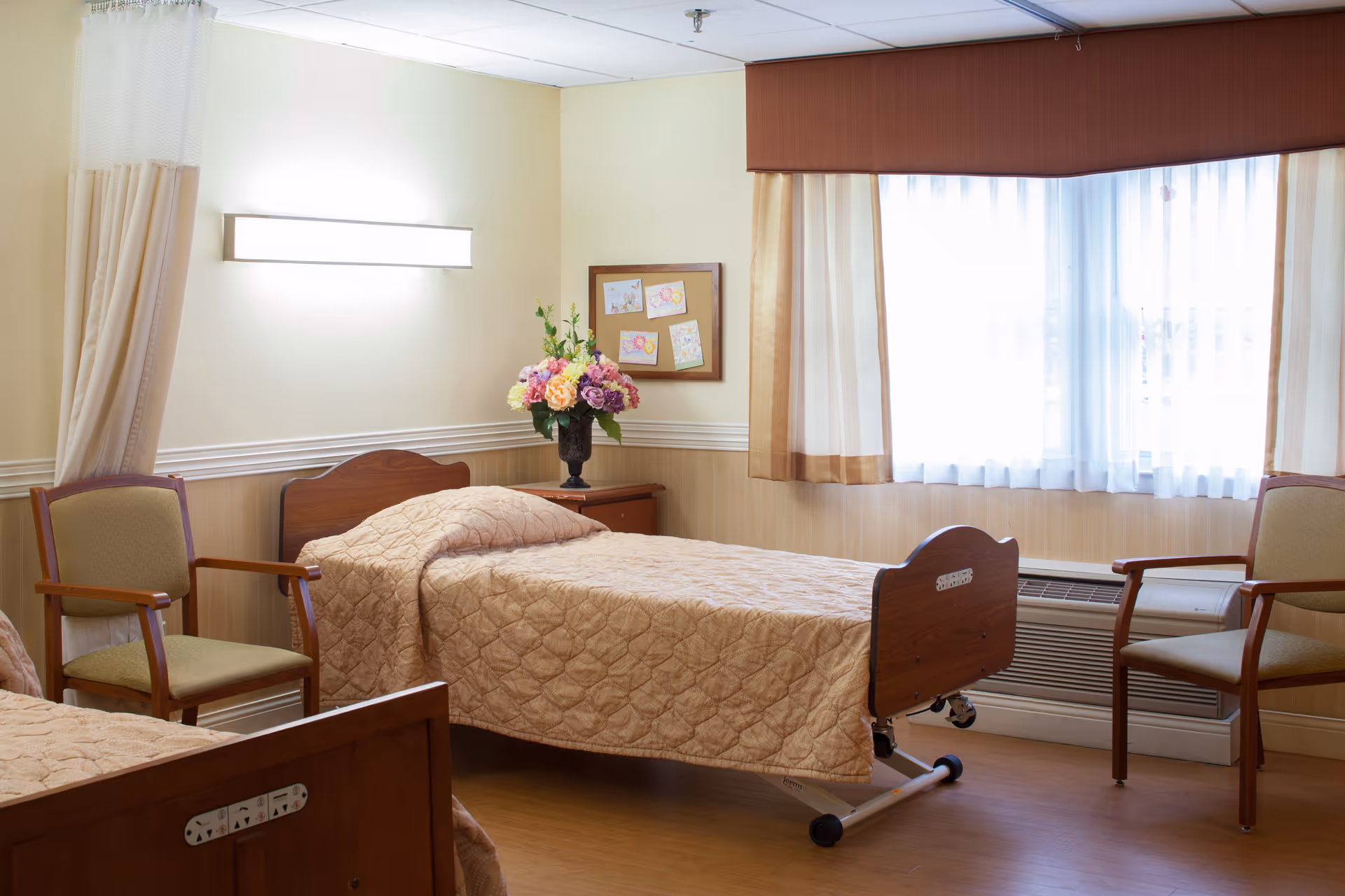 A well-lit senior living bedroom with two single beds covered in beige quilted bedspreads. There are two wooden chairs with green cushions, a wooden nightstand with a vase of colorful flowers, and a window with sheer white curtains and beige drapes. The walls are light-colored with a wooden wainscot, and a bulletin board with children's drawings is mounted on the wall.