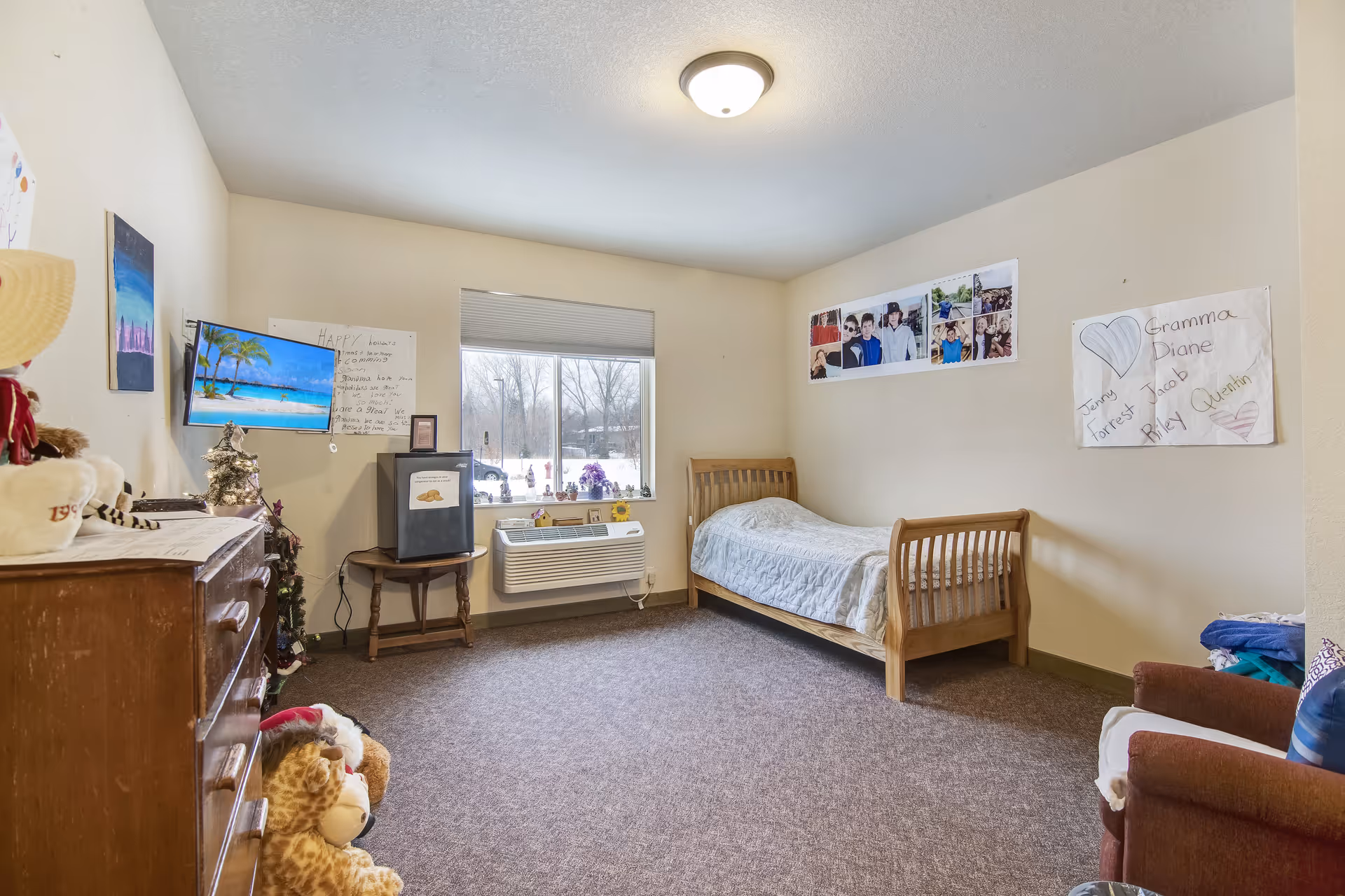 A simple bedroom in a senior living facility with a single wooden bed covered with a light-colored quilt, a wooden dresser with stuffed animals on the floor beside it, a small TV mounted on the wall, a mini refrigerator on a small table under a window, and various personal photos and handwritten notes decorating the walls.