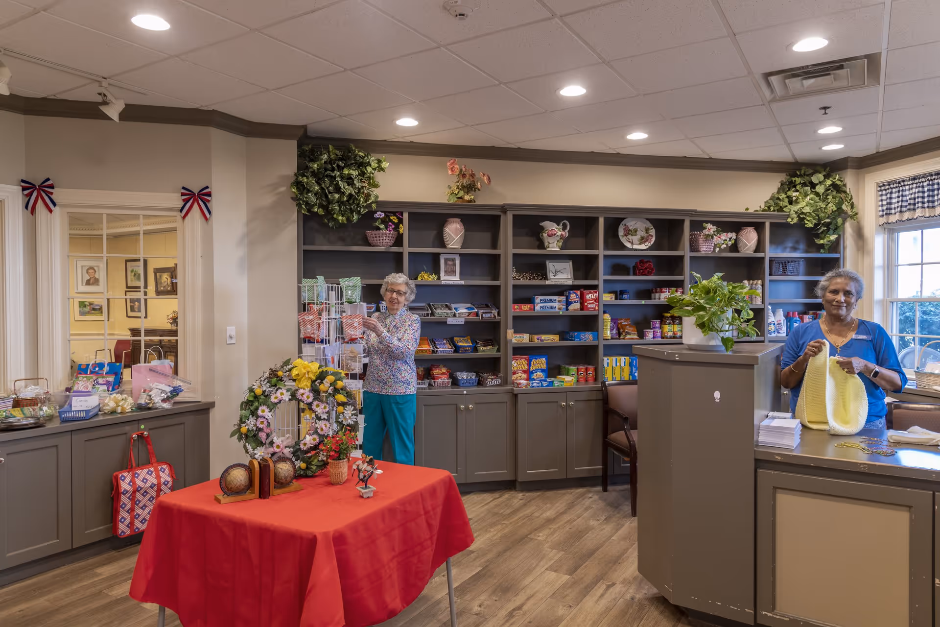 Interior of a common area in Coburg Village featuring two elderly women. One woman is standing near shelves stocked with snacks and household items, arranging greeting cards on a rotating display. The other woman is standing behind a counter, knitting a yellow garment. The room has wooden flooring, a red tablecloth-covered table with decorative items, and plants on top of the shelves and counter. There are windows with blue checkered curtains letting in natural light.