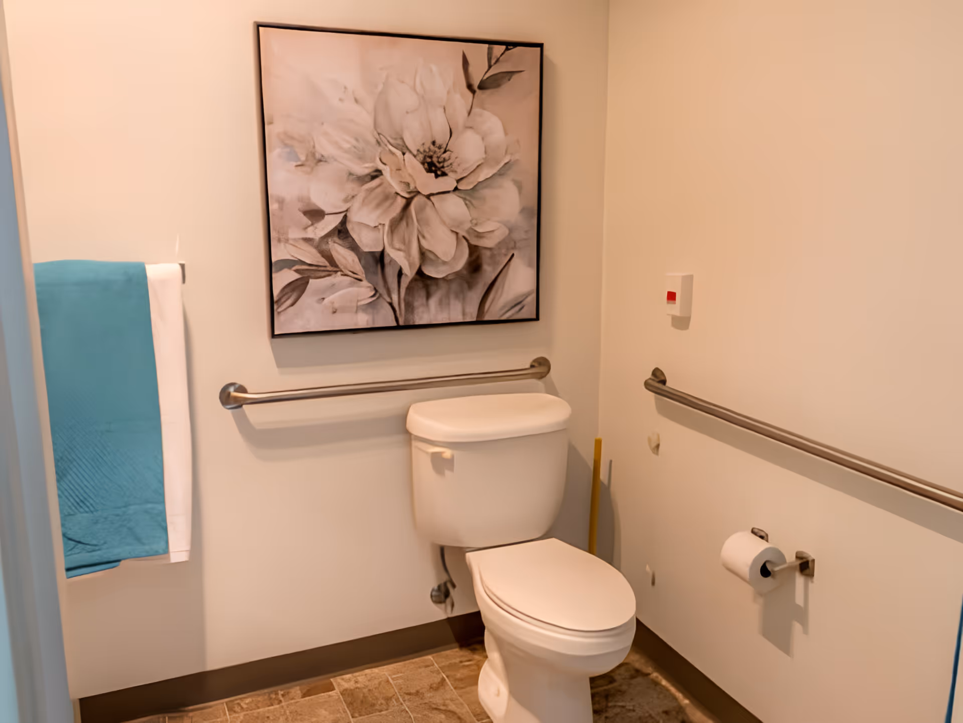 A clean and accessible bathroom with a white toilet, stainless steel grab bars on the walls, a blue and white towel hanging on a towel rack, and a large framed black and white floral artwork above the toilet.