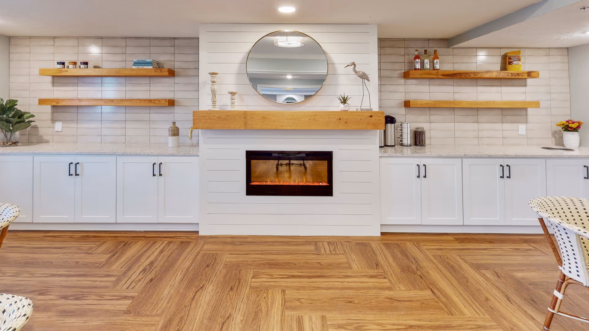 A cozy interior space featuring a modern electric fireplace with a wooden mantel, a round mirror above it, and white cabinetry with black handles on either side. Floating wooden shelves hold decorative items and bottles. The floor has a wood pattern, and part of a chair with a woven seat is visible on the right.