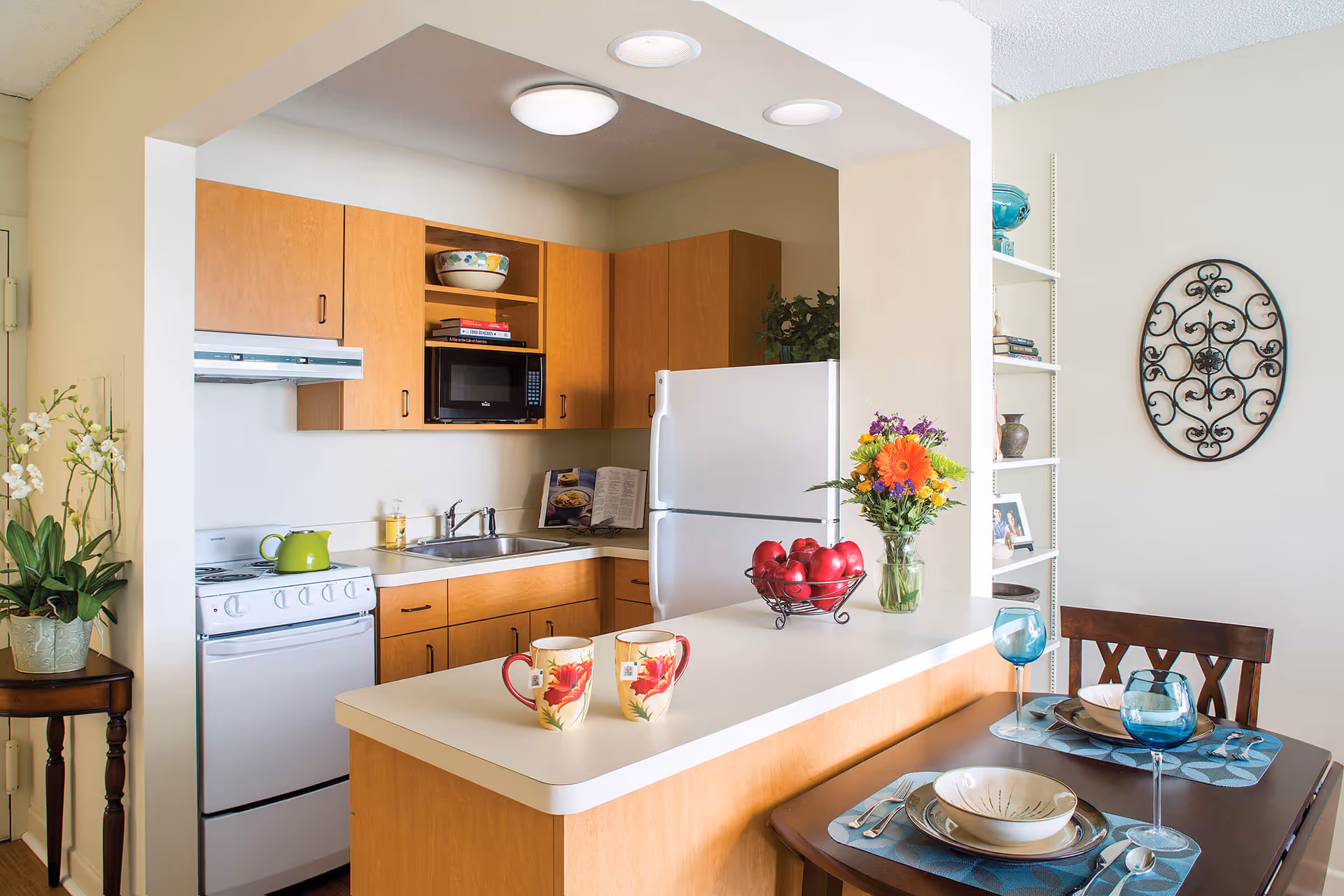 A cozy kitchen area with wooden cabinets, a white refrigerator, a stove with a green kettle, and a microwave. A countertop extends into a small dining area with two mugs, a bowl of red apples, and a vase of colorful flowers. A small dining table is set with plates, bowls, utensils, and blue wine glasses. Decorative elements include a potted plant on a small table, a wall-mounted ornamental piece, and a white shelving unit with various items.