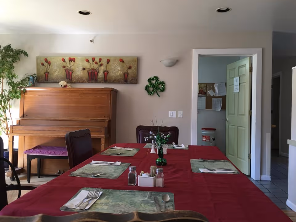 Dining room with a long table set with placemats and utensils, a piano against the wall, and an open doorway.