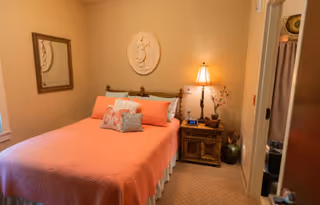 A cozy bedroom with a bed covered in a coral-colored quilt and several decorative pillows. There is a wooden nightstand with a lamp, a clock, and a small plant. A framed mirror hangs on the wall to the left, and a decorative wall plaque is centered above the bed. The room has beige walls and carpeted flooring.