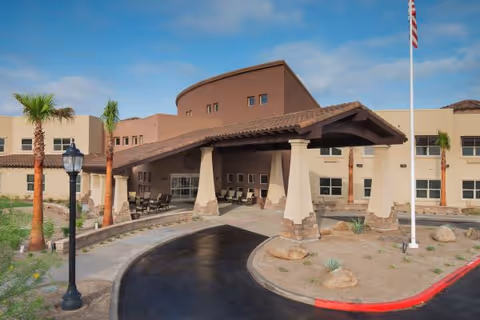 Exterior view of Solana at The Park Assisted Living Community building with a covered entrance, palm trees, a flagpole with an American flag, and a curved driveway under a partly cloudy sky.