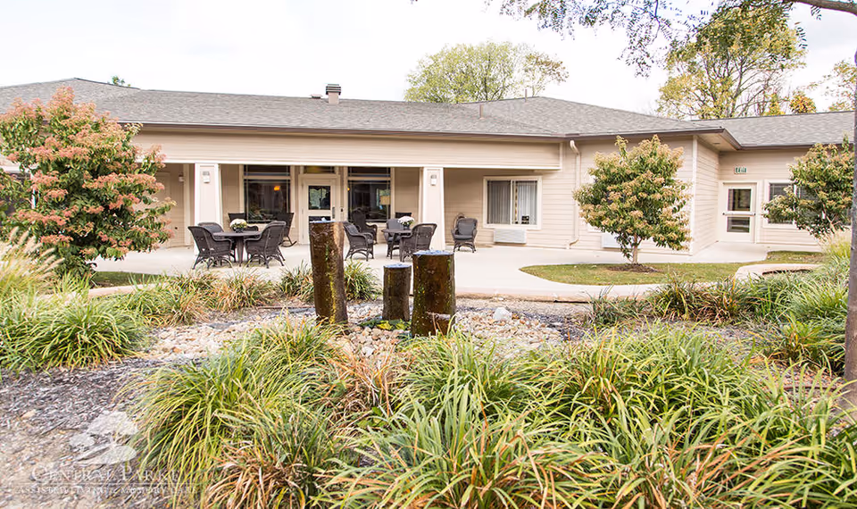 Outdoor patio area of Central Parke Assisted Living & Memory Care with several tables and chairs arranged for seating. The patio is surrounded by greenery, including bushes and small trees, and features a decorative water fountain with three vertical stone pillars in the foreground.
