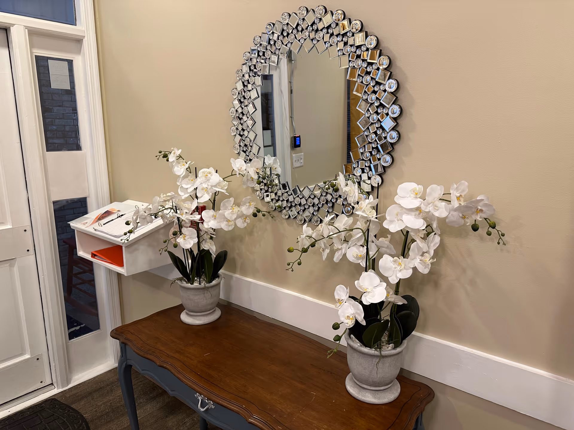 A decorative interior corner featuring a wooden table with two potted white orchid plants. Above the table is a large, ornate round mirror with a geometric frame. To the left, there is a white wall-mounted organizer with papers and a pen, next to a door with glass panels.