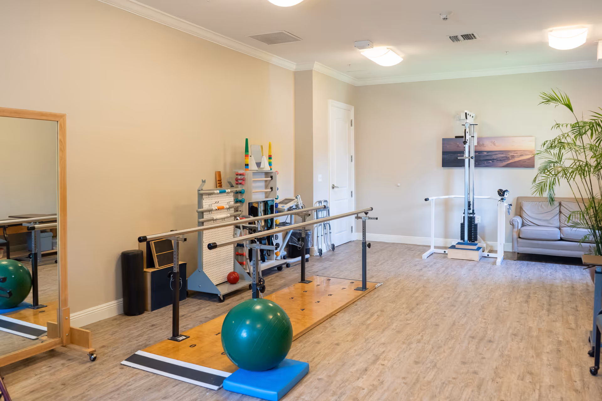 An interior therapy/exercise room with parallel bars, a green exercise ball, therapy equipment, a couch and a large mirror.