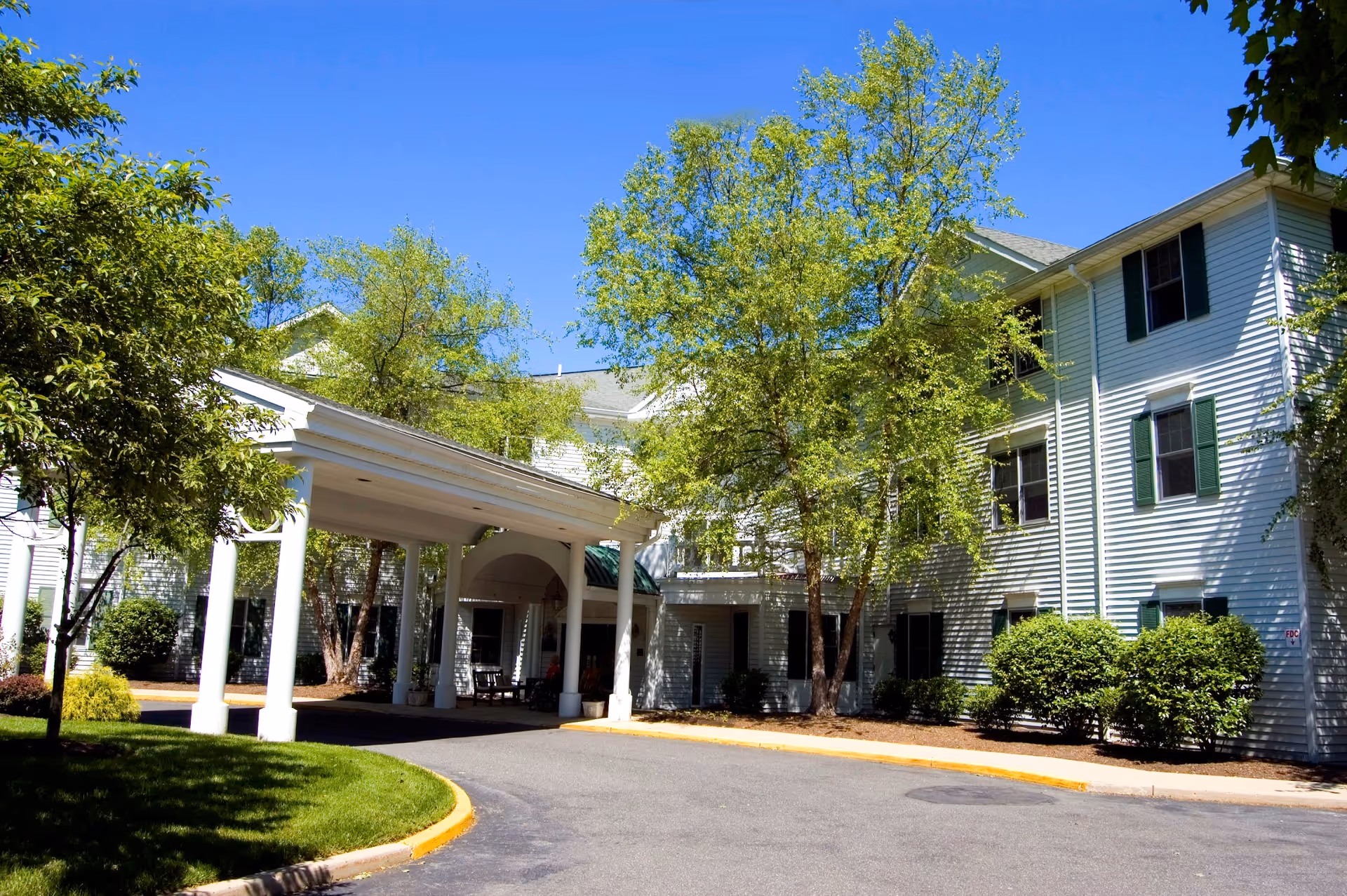 Exterior view of a white multi-story assisted living facility building with green shutters, surrounded by trees and bushes under a clear blue sky. There is a covered entrance with white columns and a driveway leading up to it.