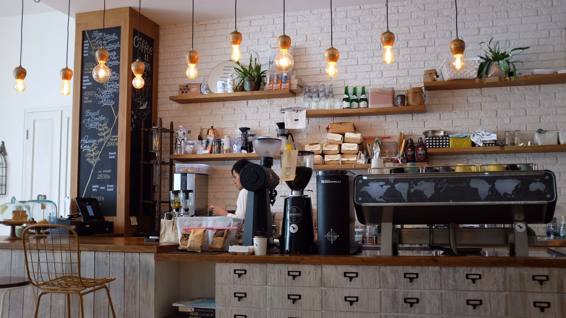 Interior view of a coffee shop counter with hanging light bulbs, a chalkboard menu, coffee machines, and shelves with various items including plants and bottles. A person is working behind the counter.