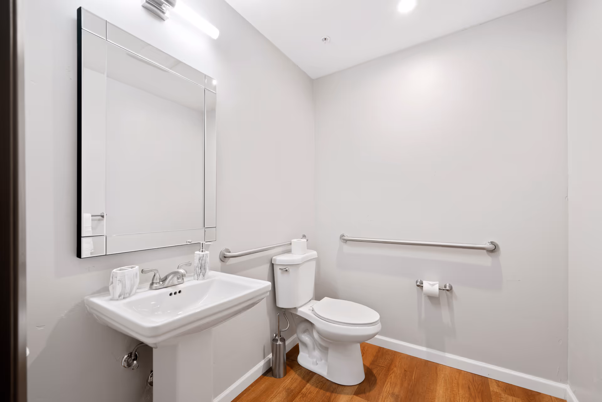 A clean and simple bathroom with a white pedestal sink, a large rectangular mirror above it, a white toilet with a roll of toilet paper on top, wooden flooring, and two metal grab bars on the walls for support.