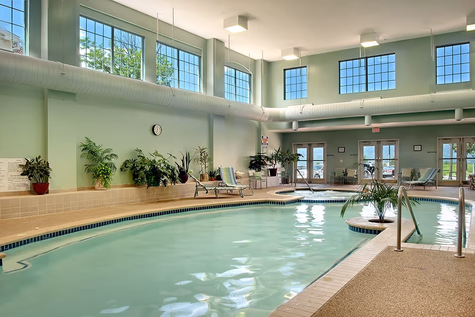 Indoor swimming pool area with light green walls, large windows letting in natural light, several potted plants along the wall, lounge chairs, and a small round hot tub adjacent to the pool.