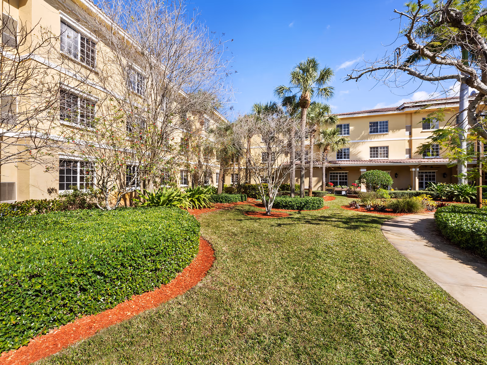Landscaped courtyard with a walkway and palm trees in front of a three-story beige building under a blue sky.