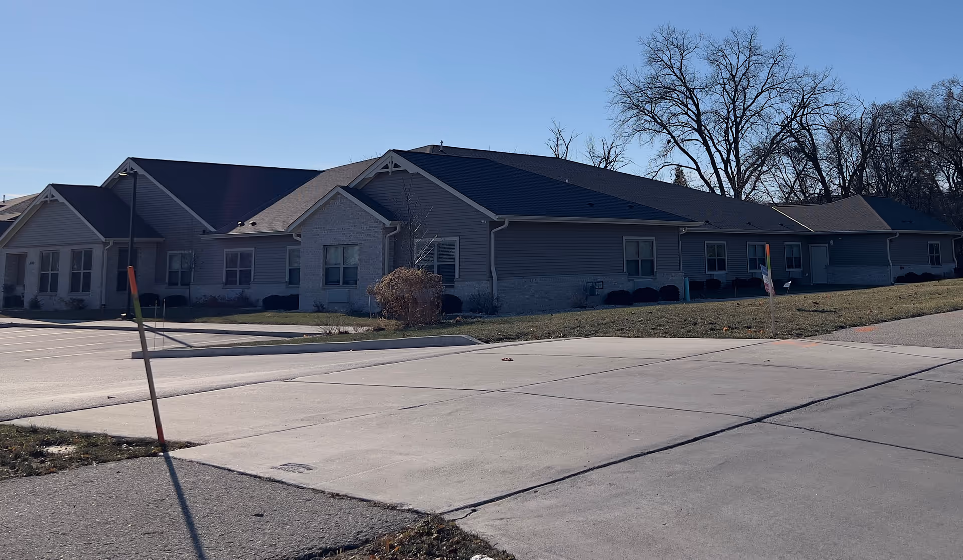 Exterior view of a single-story assisted living facility building with gray siding and brick accents, surrounded by a lawn and trees under a clear blue sky.