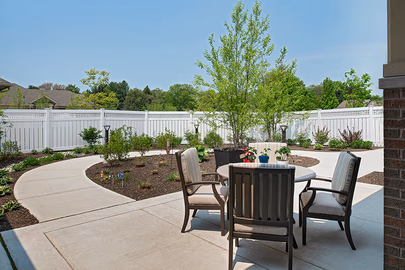 Outdoor patio area with a round table and four cushioned chairs on a concrete surface. The patio is surrounded by a garden with small plants and trees, enclosed by a white fence. The sky is clear and blue.