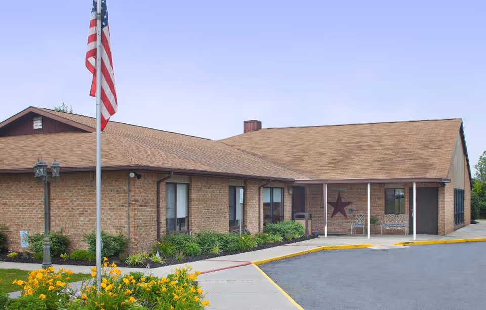 Front of a single-story brick senior living facility with an American flag, flowerbeds, and a covered entrance.