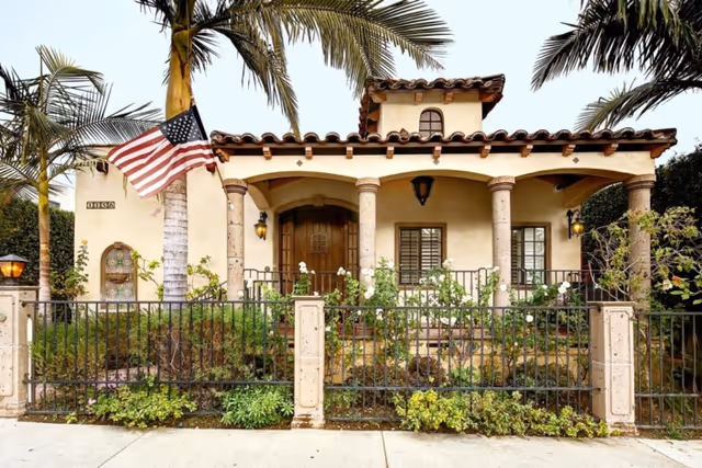 Front exterior view of a single-story house with a tiled roof, arched porch supported by columns, an American flag mounted on a palm tree, and a black metal fence with greenery and flowers in front.