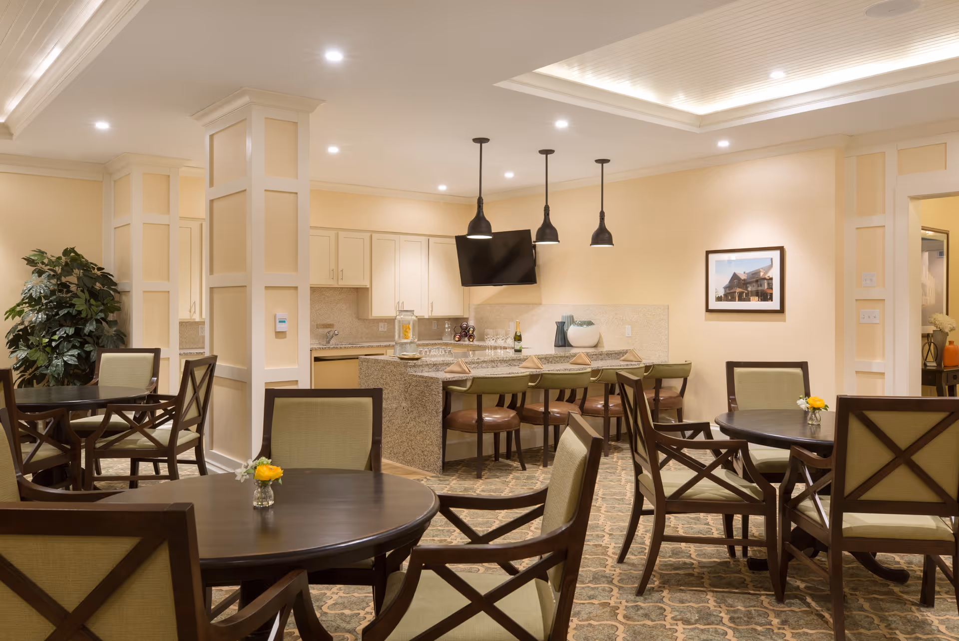 A well-lit dining area with round wooden tables and cushioned chairs arranged around them. In the background, there is a kitchen area with a granite countertop island, bar stools, pendant lights hanging from the ceiling, and a wall-mounted TV. The room has beige walls, decorative columns, and a patterned carpet. A small vase with yellow flowers is placed on each table.