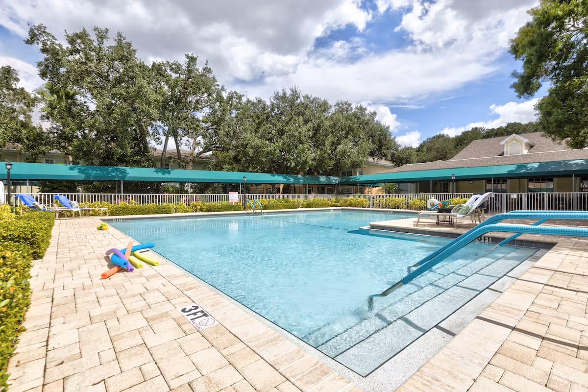 Outdoor swimming pool with clear blue water surrounded by a tiled deck. Poolside chairs with cushions are arranged on a raised platform. Trees and a building with a green awning are visible in the background under a partly cloudy sky. Colorful pool noodles are placed on the pool deck near the edge.