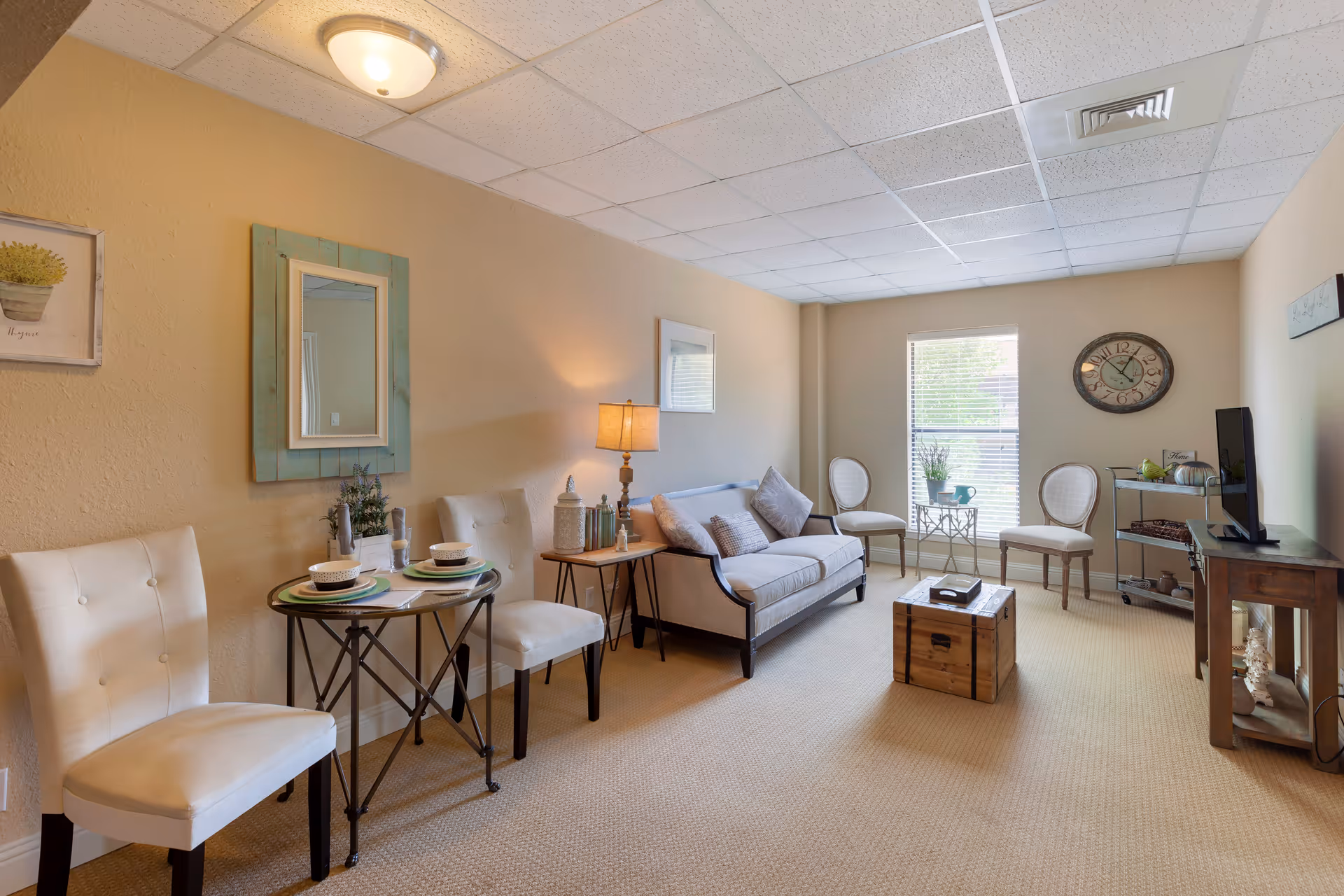 A cozy living room in an assisted living facility featuring a beige sofa with cushions, two white chairs around a small round table set with plates and bowls, a wooden chest used as a coffee table, two additional chairs near a window with blinds, a wall clock, a TV on a wooden stand, and soft lighting from a table lamp.