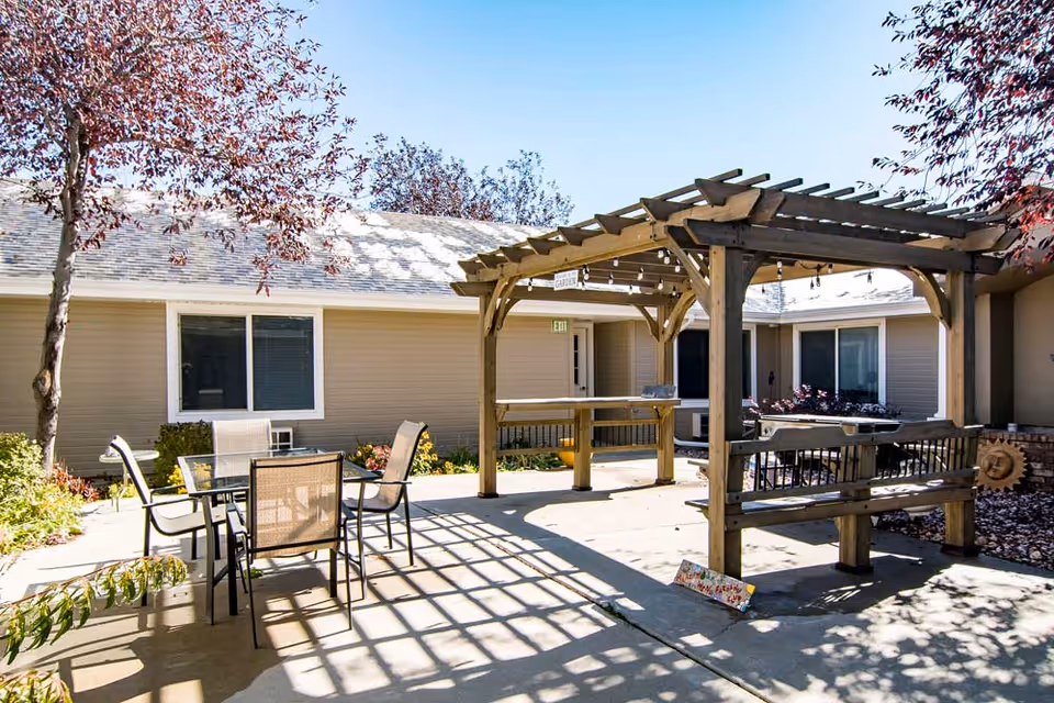 Sunny courtyard featuring a wooden pergola, outdoor table and chairs, and surrounding single-story building windows.
